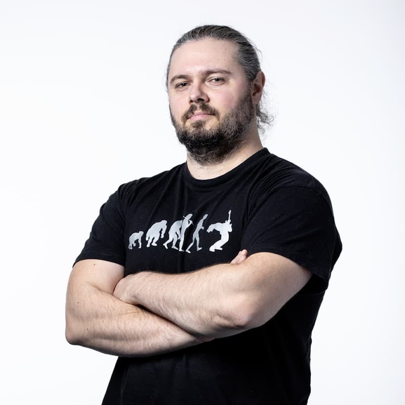 Portrait with arms crossed: beard and hair tied back, black crew-neck t-shirt with evolution graphic, white studio background.